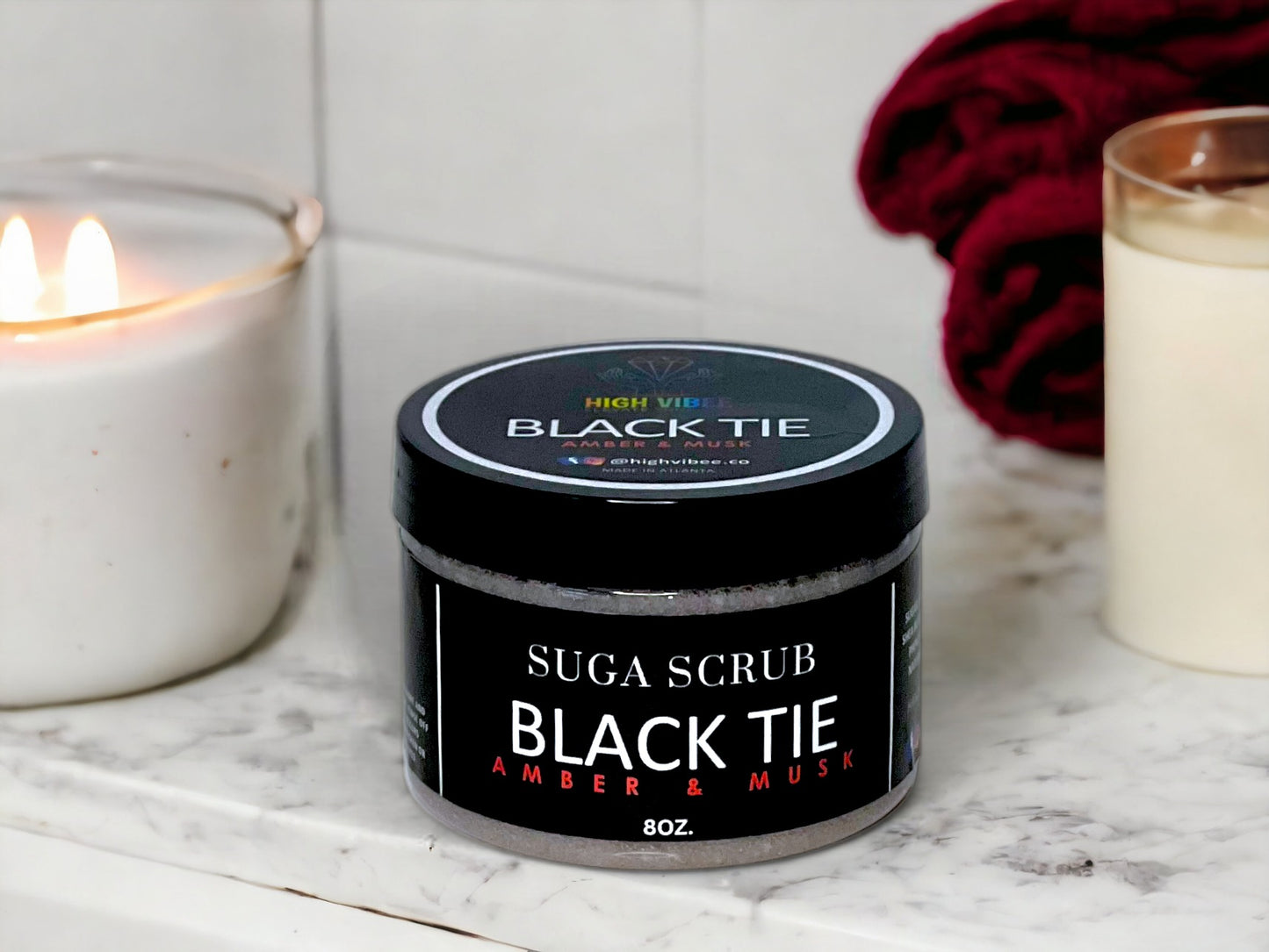 In this photo, there is a jar of High Vibee's "Black Tie" Suga Scrub scented with Amber & Musk. In the photo it is sitting next to a lit white candle.