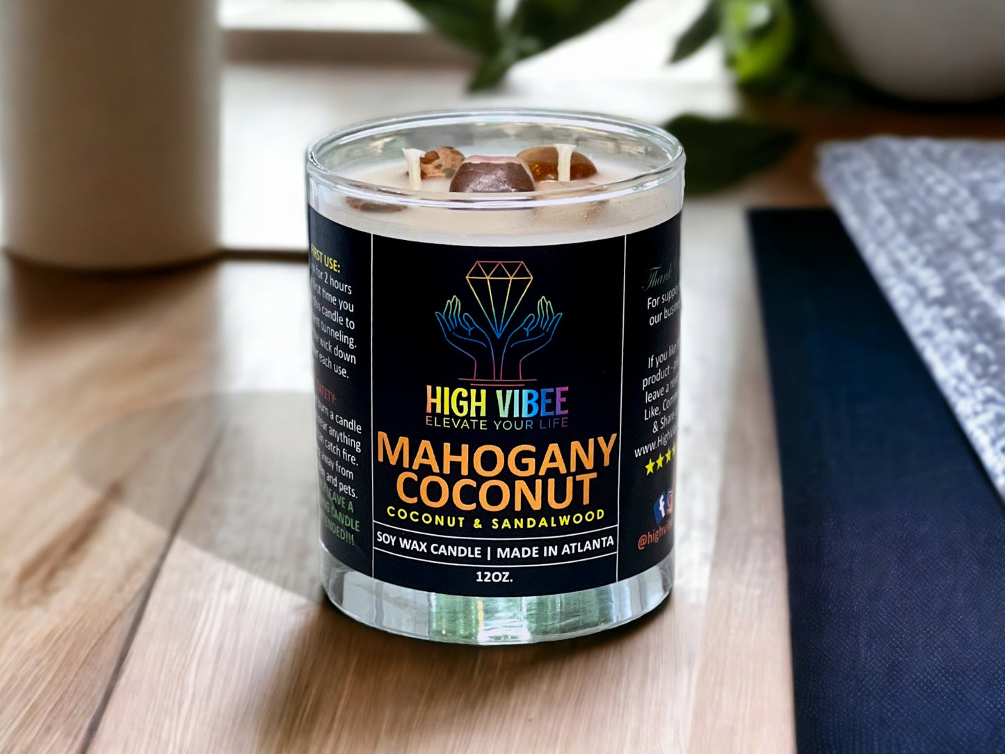 In this photo, there is a jar of High Vibee's “Mahogany Coconut” 12oz. Candle, scented with Coconut & Sandalwood. In the photo, the top is off of the candle, and there are two wicks along with brown crystals inside.