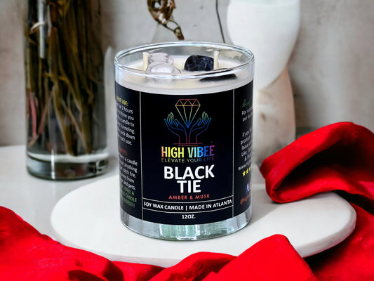 In this photo, there is a jar of High Vibee's "Black Tie" 12oz. Candle, scented with Amber & Musk. In the photo, the top is off of the candle, and there are two wicks along with black & white crystals inside.