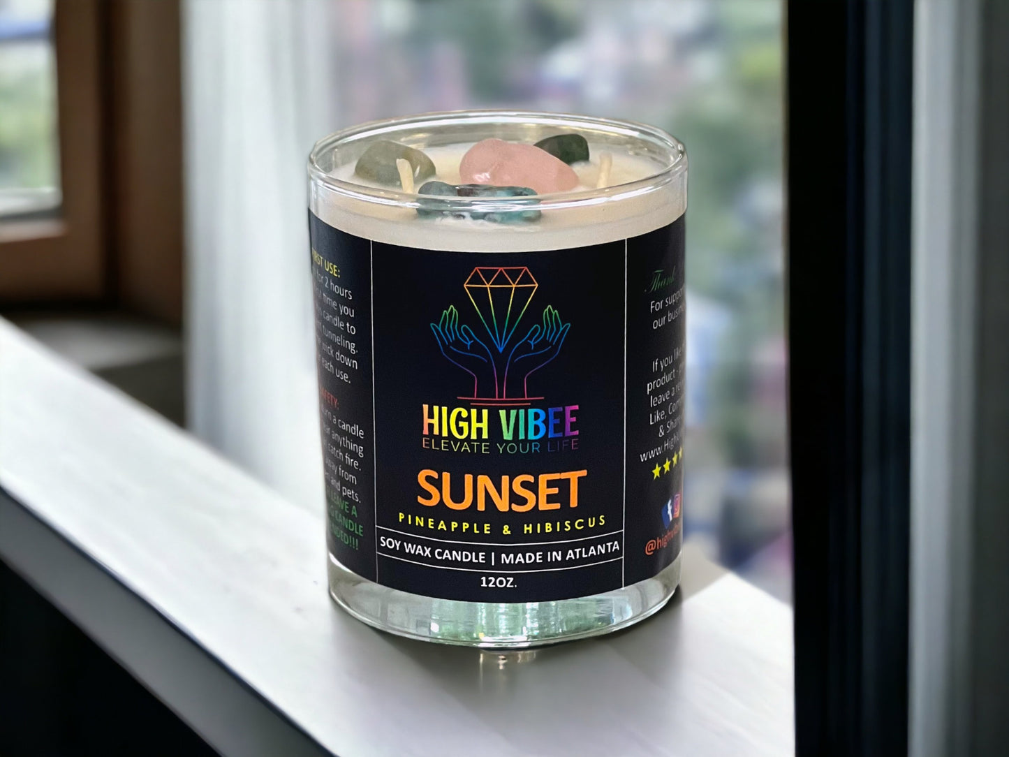 In this photo, there is a jar of High Vibee's “Sunset” 12oz. Candle, scented with Pineapple & Hibiscus. In the photo, the top is off of the candle, and there are two wicks along with pink, blue and green crystals inside.