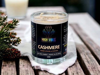 In this photo, there is a jar of High Vibee's “Cashmere” 12oz. Candle, scented with Warm Spices & Cocoa. In the photo, the top is off of the candle, and there are two wicks along with clear crystals inside.